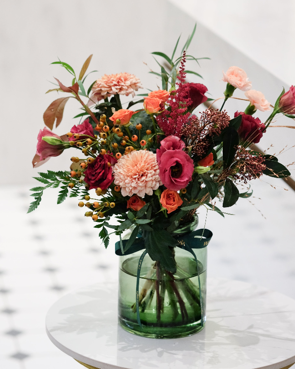 A vibrant floral arrangement in a clear green glass vase, featuring pink, red, and peach flowers with green foliage, displayed on a white marble-topped table against a light background.