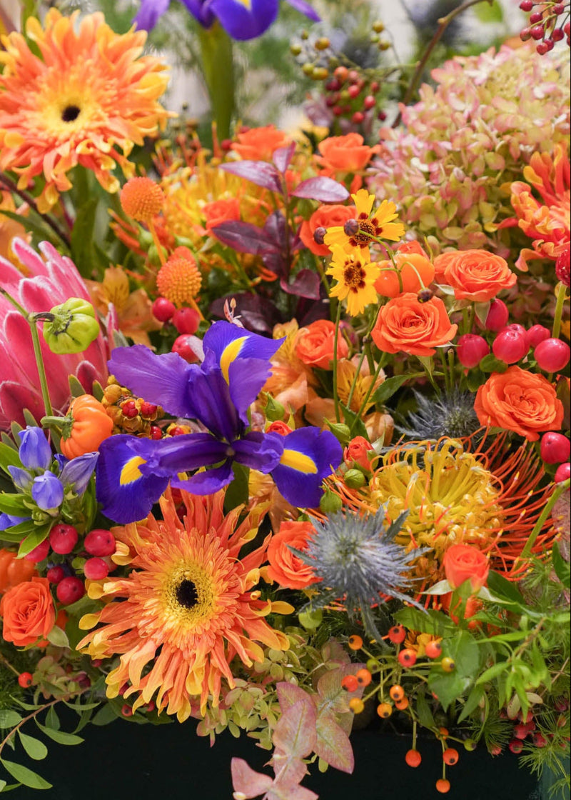 A vibrant, dense floral arrangement featuring orange gerbera daisies, purple irises, pink protea, orange roses, and green foliage in a dark green container.