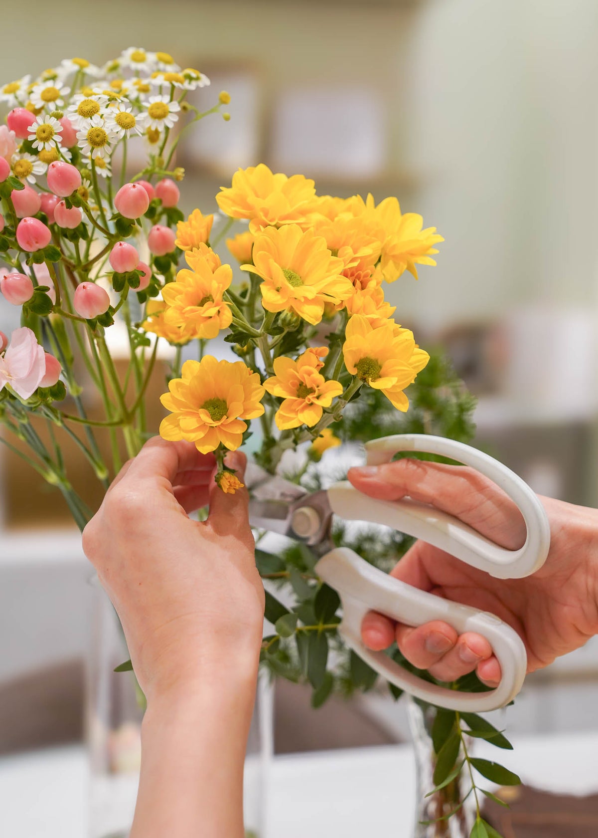 A close-up of hands using white pruning shears to trim a bouquet of yellow flowers, with pink buds and white daisies also visible in a soft-focus indoor setting.