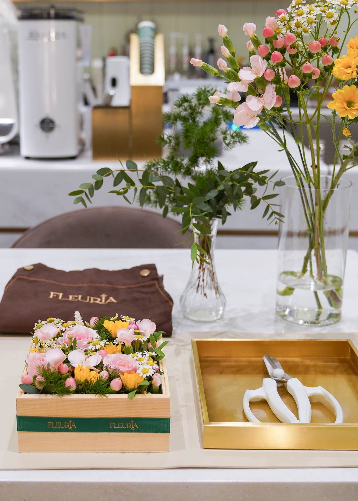 A styled floral arrangement display on a countertop, featuring a wooden box of pink, peach, and yellow flowers with a green Fleurin brand band, a brown Fleurin pouch, a gold tray with white-handled pruning shears, and two glass vases with greenery in a bright kitchen-like setting.