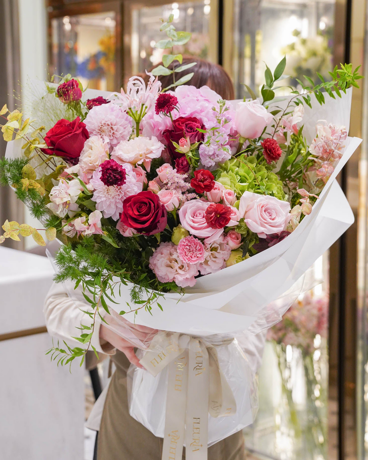 A large, vibrant bouquet of pink, red, and white flowers with green foliage, wrapped in white paper with a cream ribbon, held by a person in a beige outfit inside a shop with glass doors.