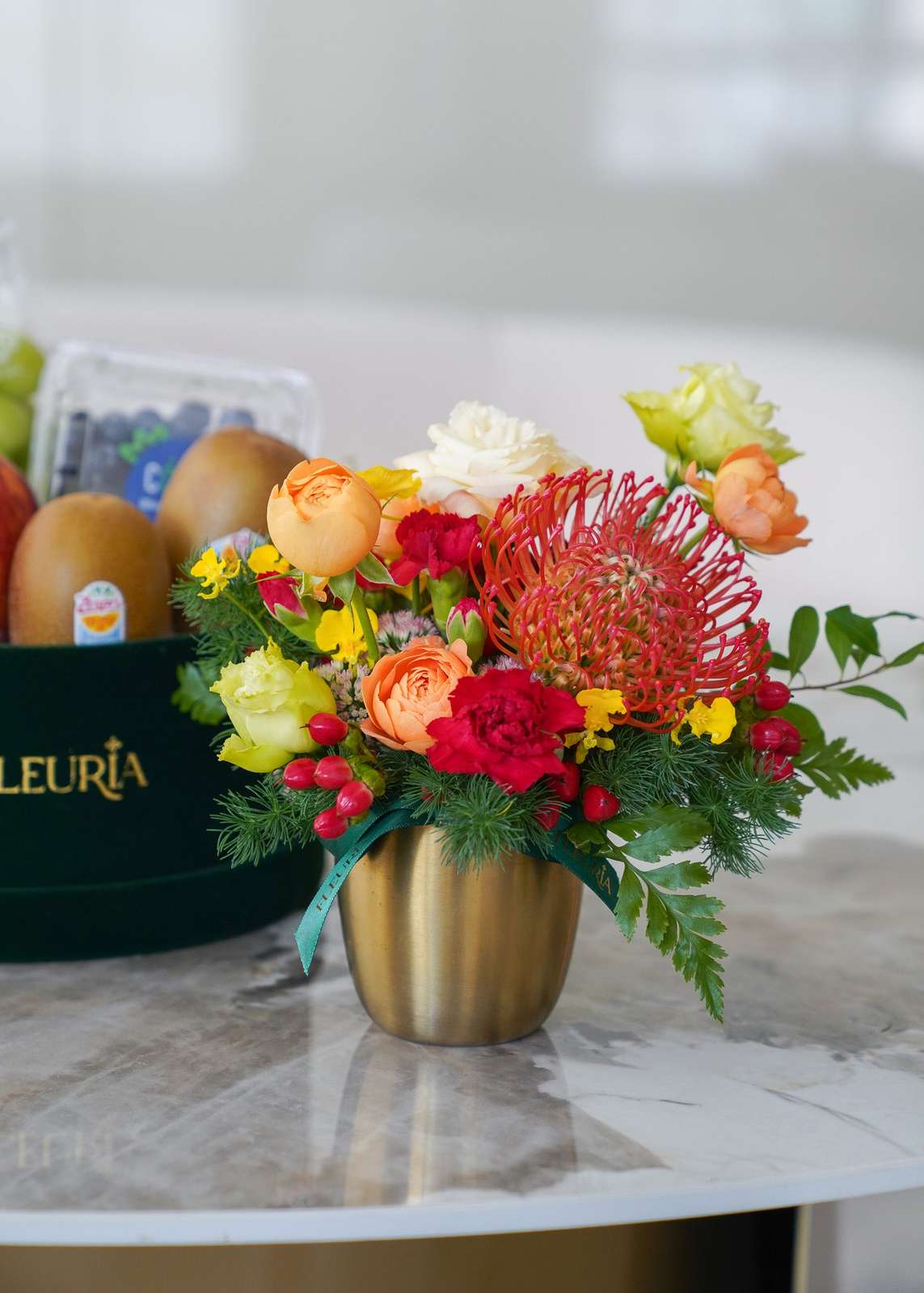 A vibrant floral arrangement in a metallic gold vase, featuring red protea, orange roses, yellow flowers, and green foliage,