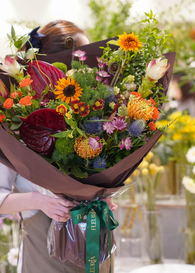 A large, vibrant mixed floral bouquet wrapped in dark brown paper and tied with a green ribbon, held by a person whose head and hands are partially visible in a bright, softly blurred indoor setting.