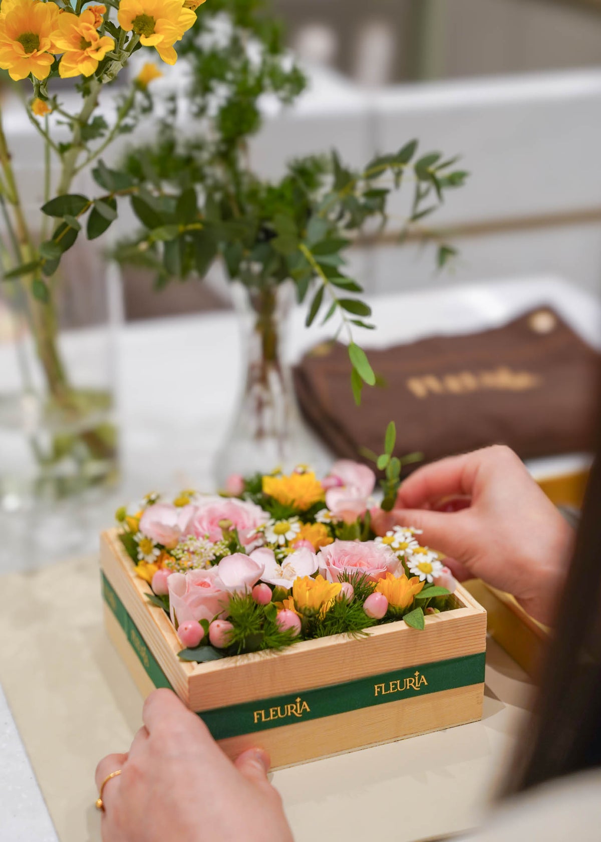 A close-up of hands arranging a wooden box floral arrangement with pink and yellow flowers and green foliage. The box has a green band with the brand name 'FLEURIA' printed in gold.