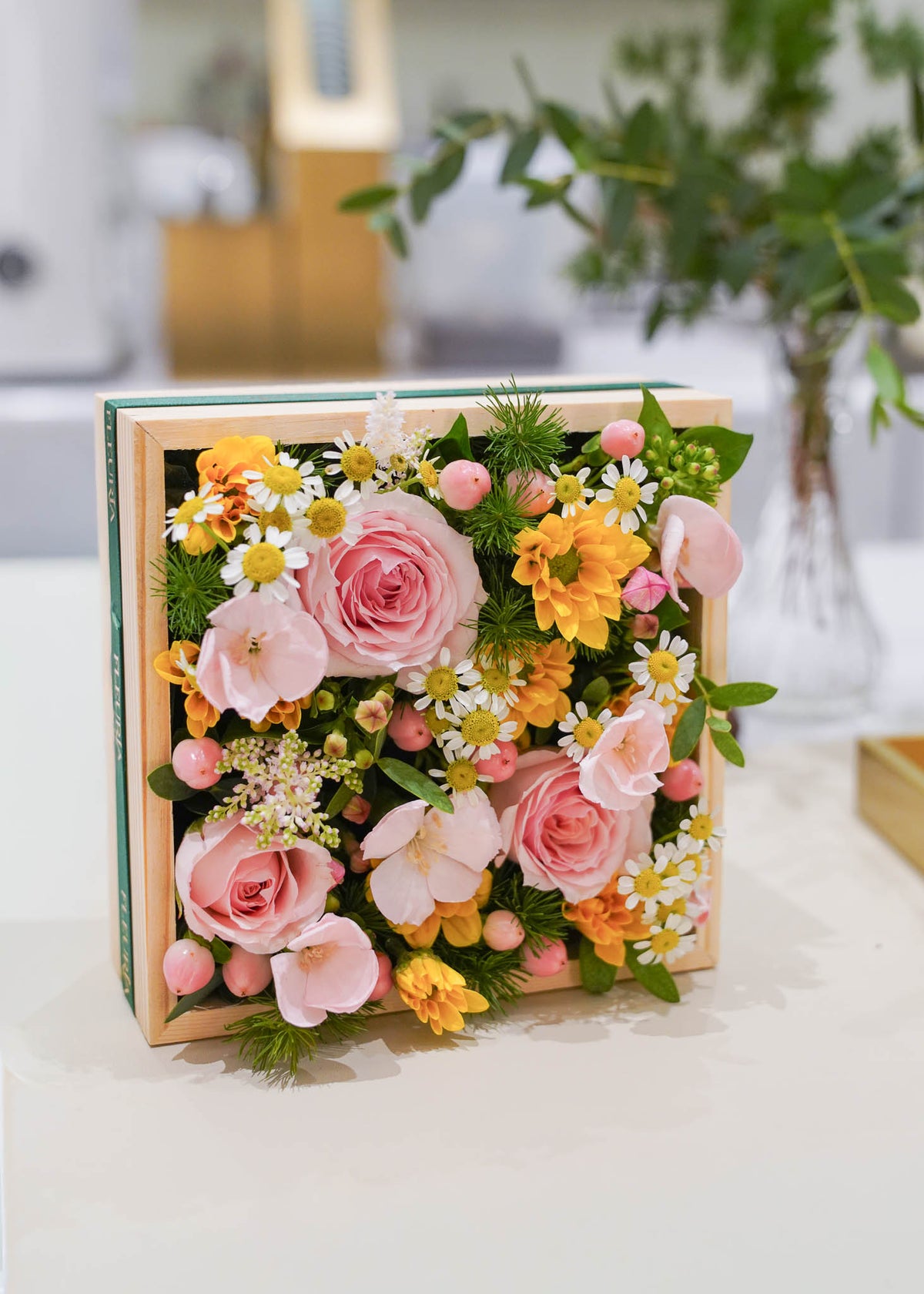A square wooden gift box filled with a colorful floral arrangement featuring pink roses, yellow daisies, white daisies, and green foliage, displayed on a light surface with a softly blurred indoor background.