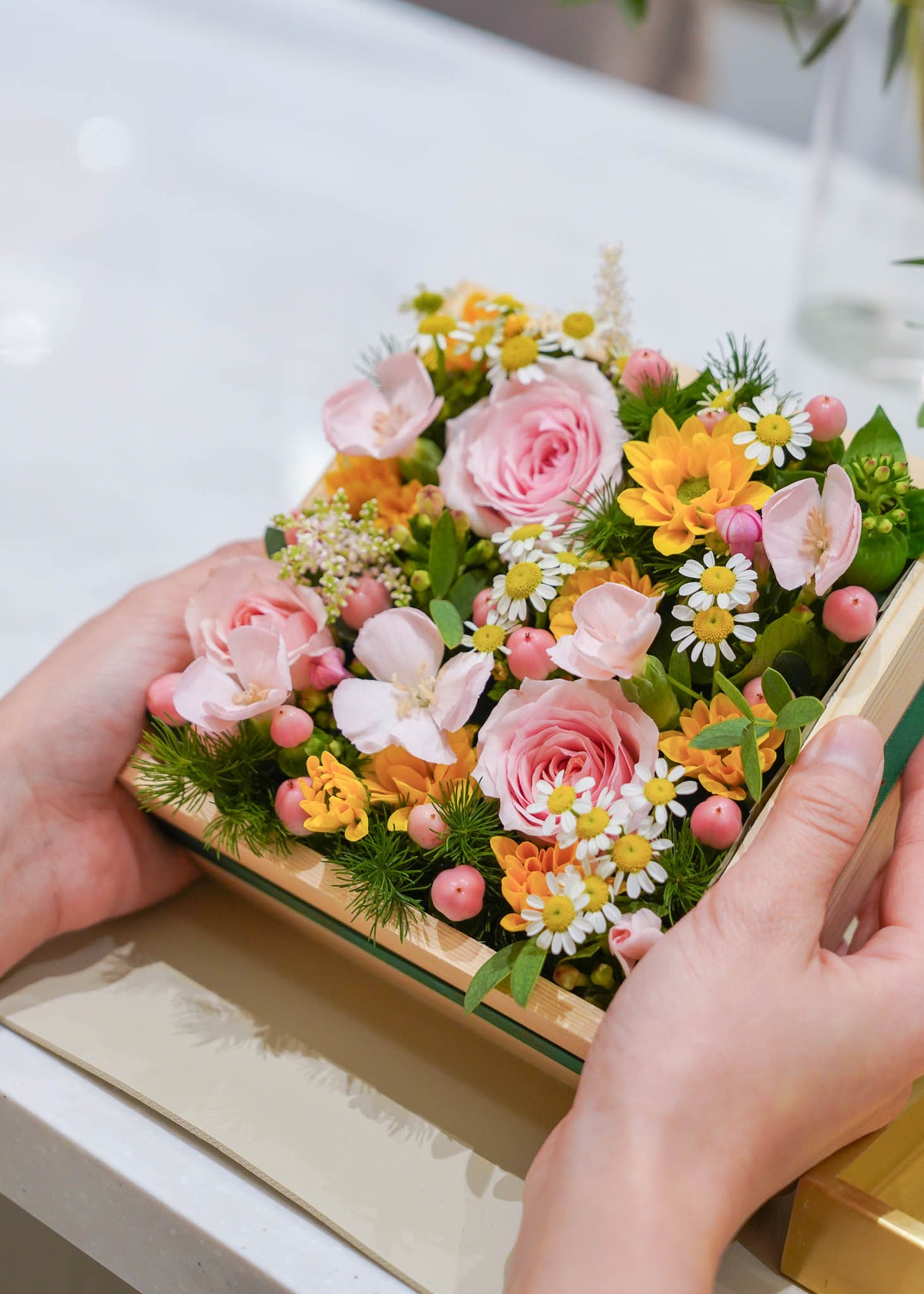 A close-up of a hand holding a square wooden box filled with a colorful floral arrangement featuring pink roses, yellow flowers, white daisies, and green foliage.