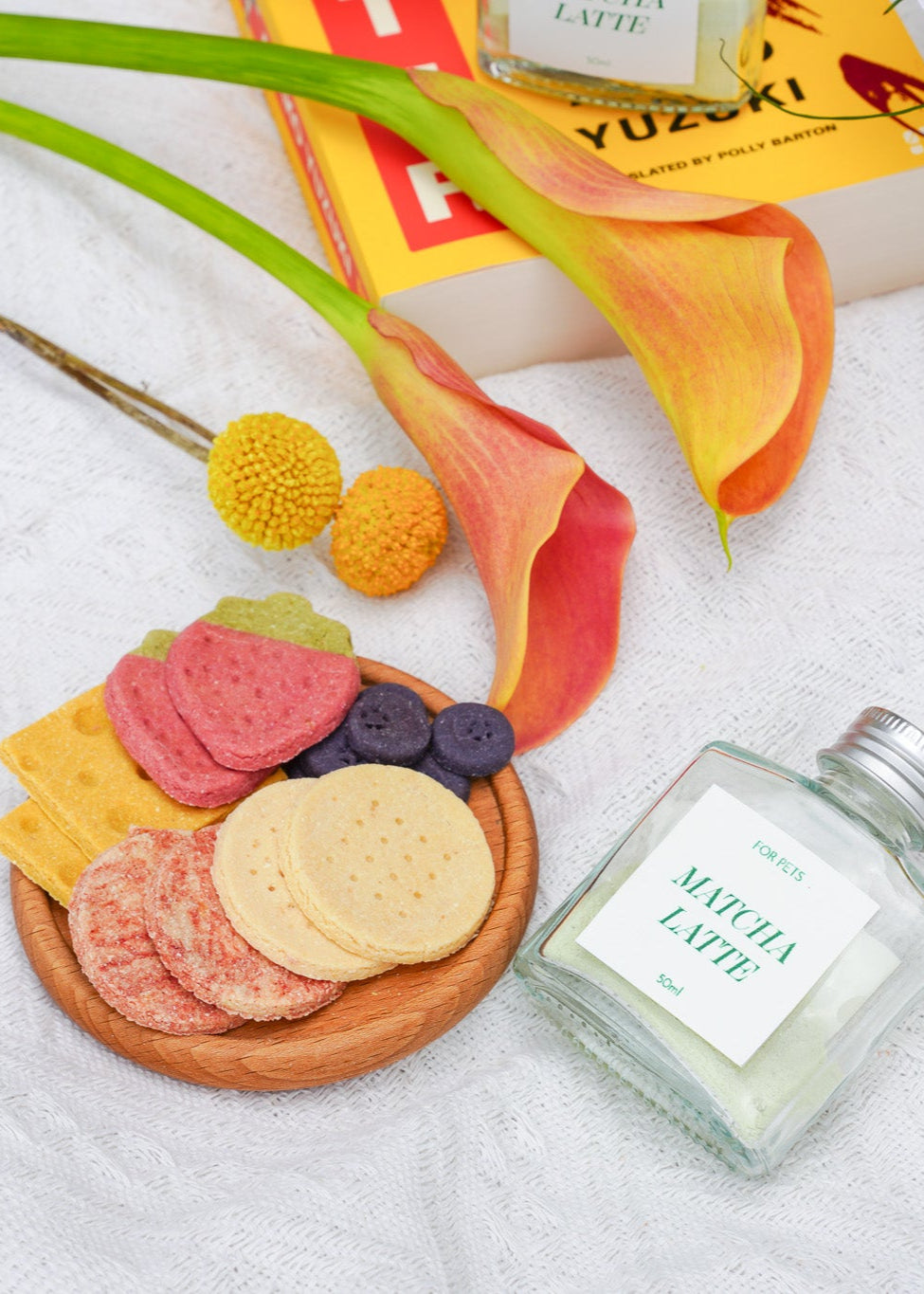 Small wooden plate with colorful cookies, a bottle labeled 'Matcha Latte', and flowers on a white blanket.