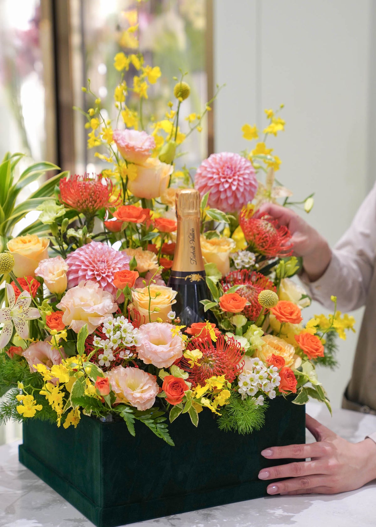 A vibrant floral arrangement in a dark green box on a table, featuring a bottle of champagne, with a person's hands adjusting the flowers in a bright indoor setting.