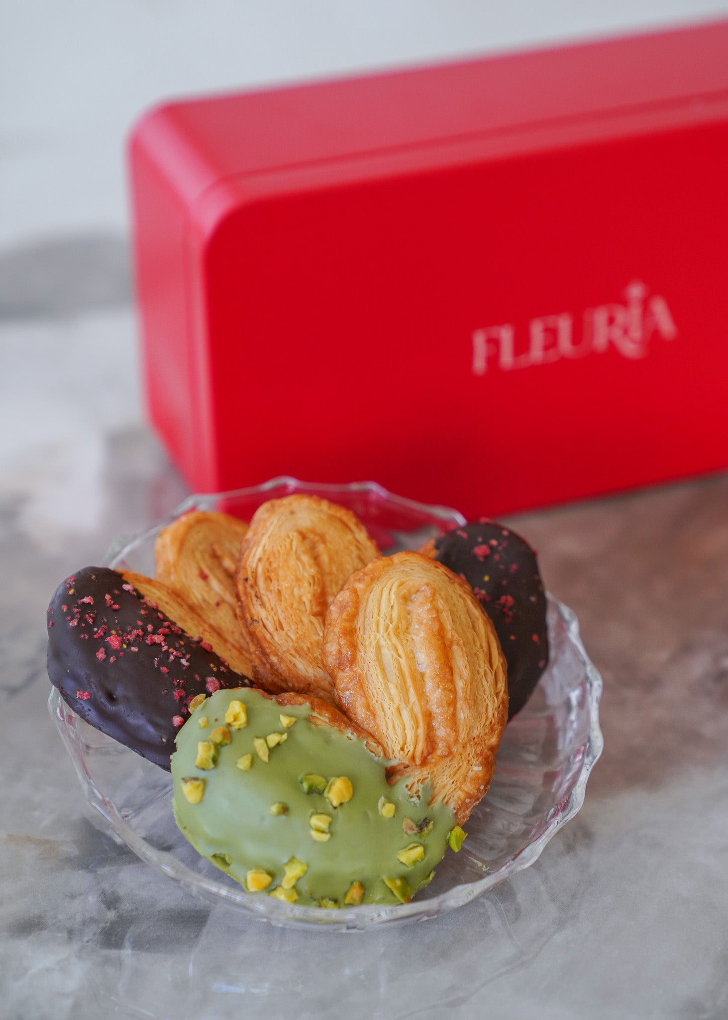 Four assorted pastries in a clear glass dish, with a red rectangular box labeled FLEURIA in the background on a light gray surface.