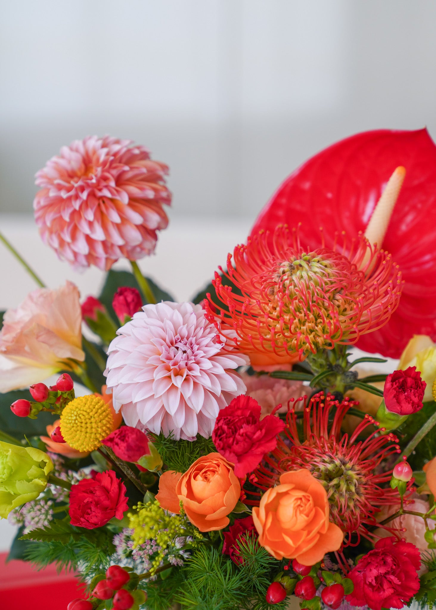 A vibrant floral arrangement featuring a variety of colorful flowers including pink and white dahlias, red protea, orange roses, red anthuriums, and green foliage.