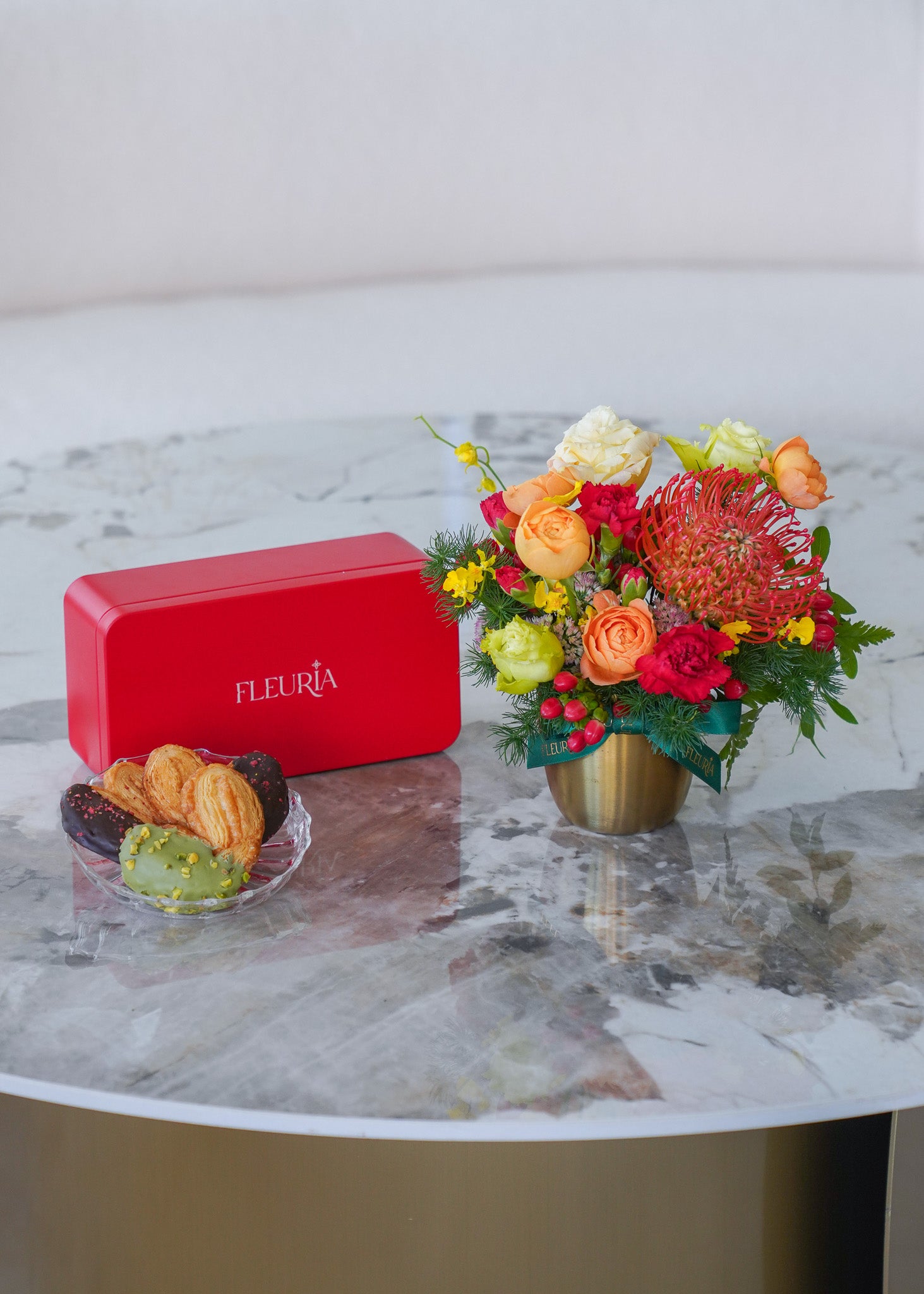 A red rectangular box with the brand name 'FLEURIA' in white lettering sits on a round white marble table next to a colorful flower arrangement in a gold vase and a small plate of pastries.