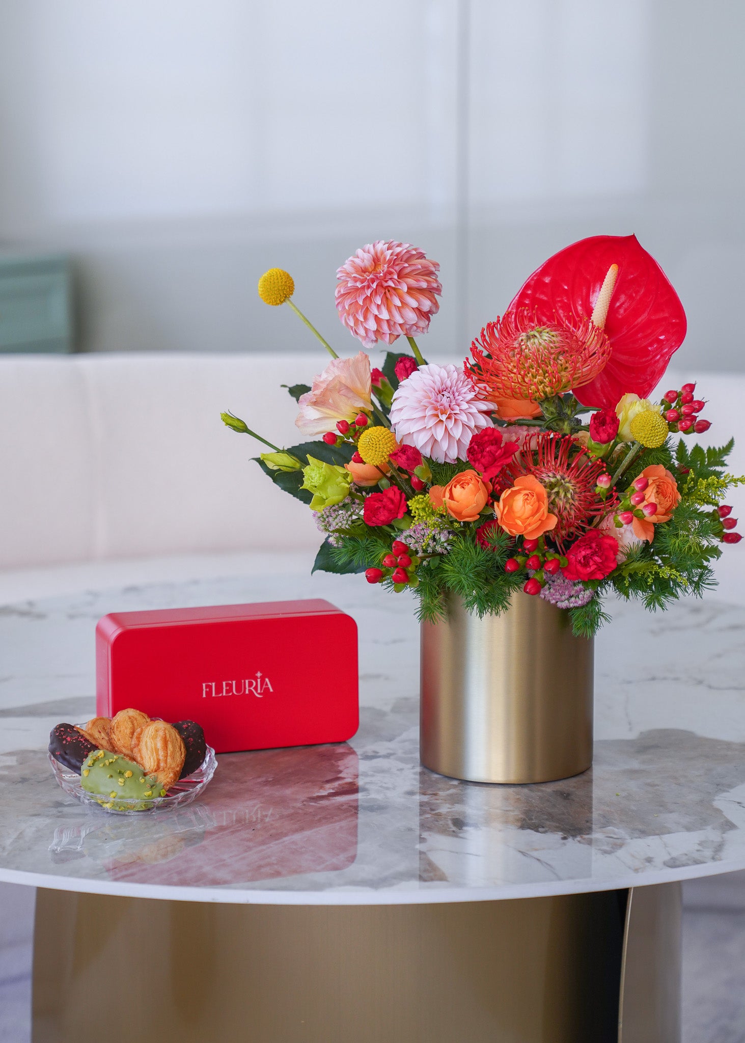 A vibrant floral arrangement in a metallic gold cylinder vase on a marble-topped table, next to a red rectangular box with the brand name 'fleuria' and a small glass dish of cookies.