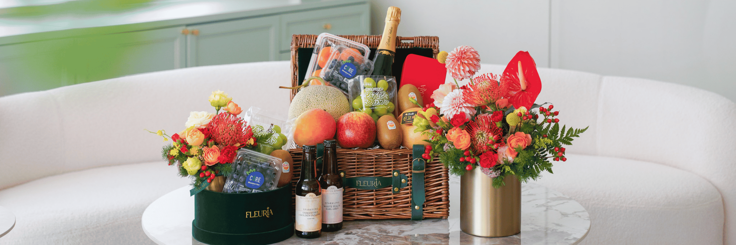 A luxury fruit and champagne gift hamper arranged on a marble-top table, flanked by two floral arrangements in round green and gold containers. The central wicker basket contains assorted fruits, a bottle of champagne, and a red gift box, with the brand name FLEURA visible on the hamper and left container.