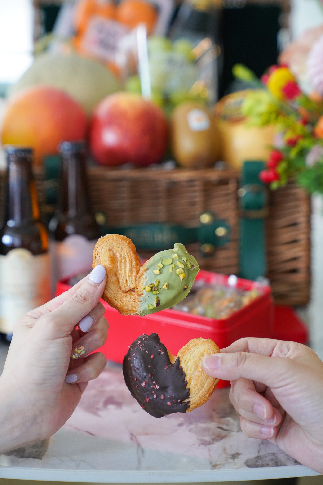 Two hands holding heart-shaped pastries dipped in chocolate and pistachio spread, with a blurred picnic-style background featuring fruit, bottles, and a basket.