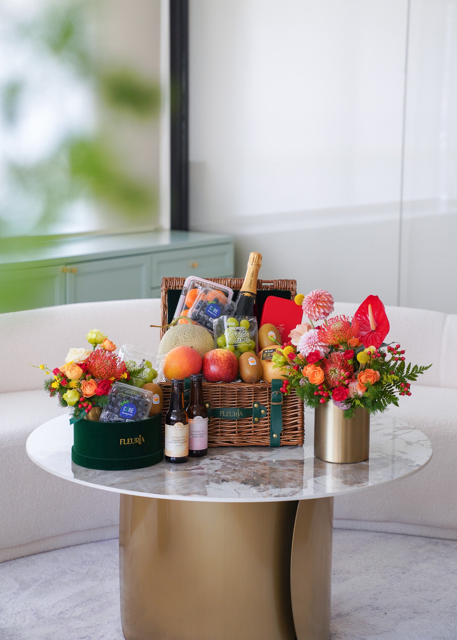 A luxury gift hamper arranged on a round marble-top table in a bright, modern room. The hamper includes a wicker basket with assorted fruits, bottles, and packaged goods, flanked by two floral arrangements in a dark green box and a gold vase.