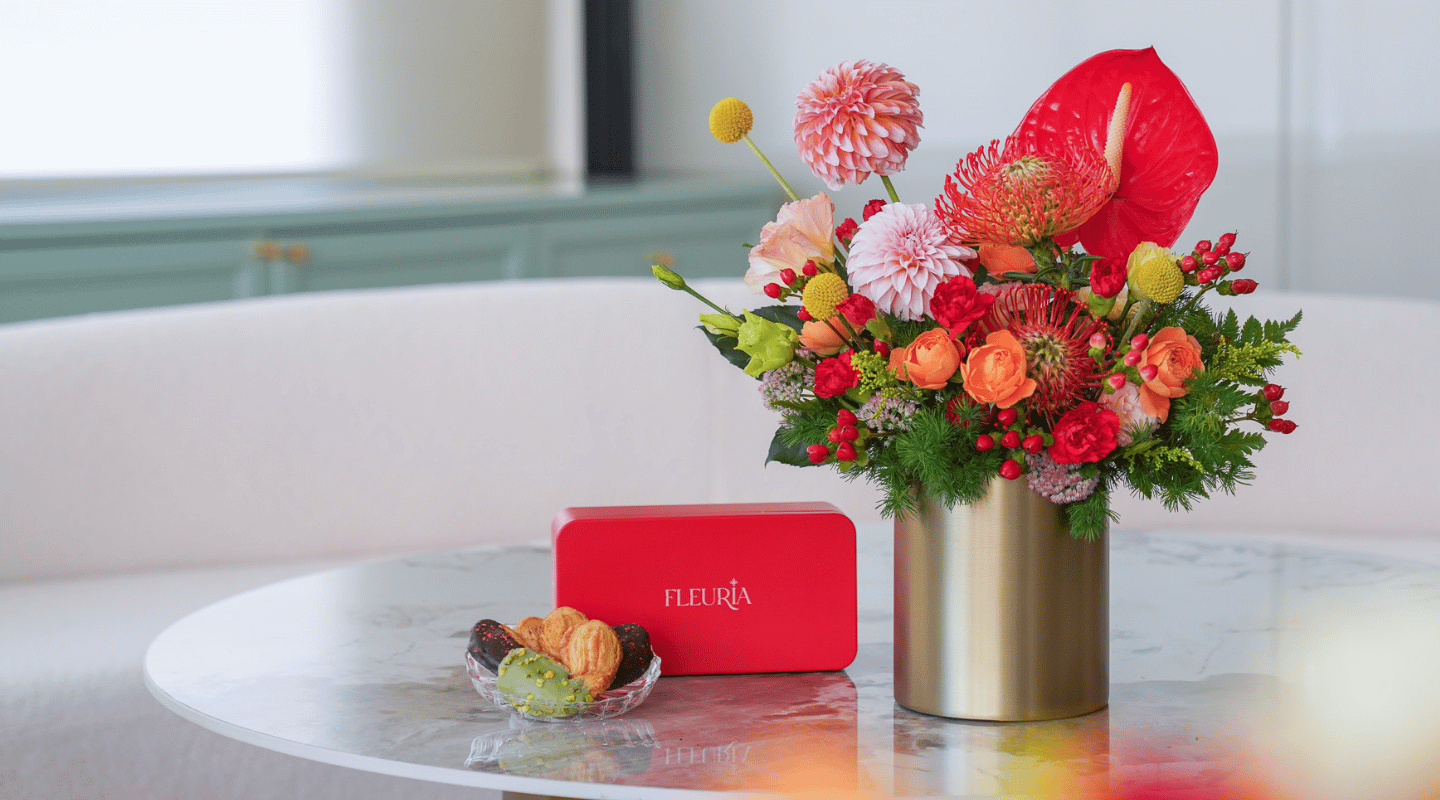 A vibrant floral arrangement in a metallic gold cylinder vase sits on a round white marble table next to a red rectangular box with the brand name 'FLEURIA' and a small glass dish of cookies, set against a softly blurred modern interior background.