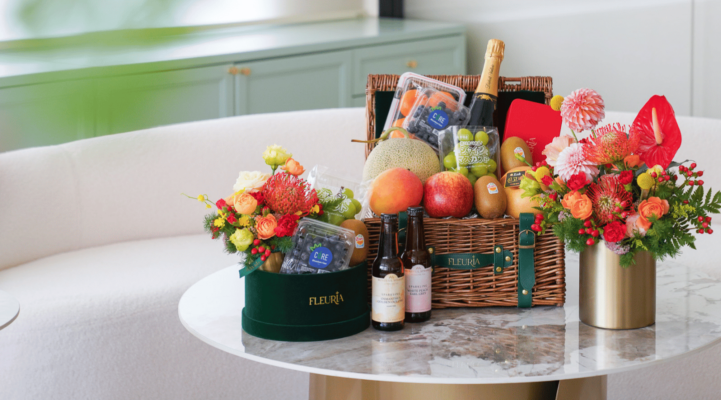 A luxury gourmet gift hamper styled on a marble-topped table in a bright, modern room. The hamper contains a bottle of champagne, assorted fruits (mango, papaya, grapes, pear), and packaged snacks, flanked by two vibrant floral arrangements in a green round box and a gold vase.