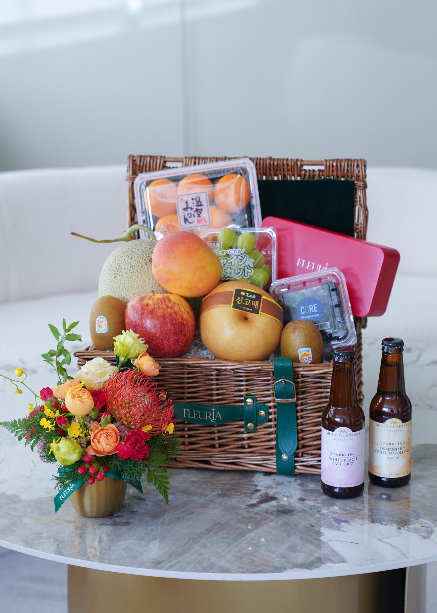 A wicker gift basket filled with assorted fruits, packaged vegetables, and a pink tin, styled on a round marble table with a small floral arrangement and two brown bottles. The basket has a green ribbon with the brand name 'FLEURA' on it.