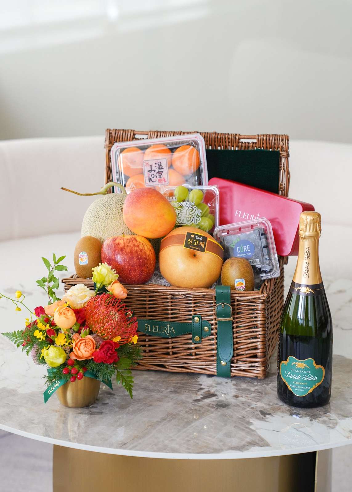 A wicker picnic basket filled with assorted fruits, a pink gift box, and packaged items, styled on a marble table with a smal