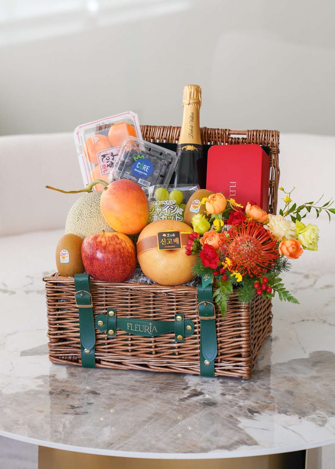 A wicker picnic basket filled with assorted fruits, a bottle of champagne, a red gift box, and a colorful flower arrangement,