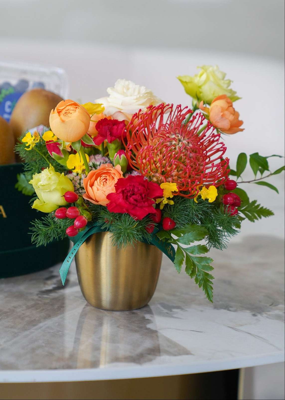 A vibrant floral arrangement in a metallic gold vase, featuring red protea, orange roses, yellow flowers, and green foliage,