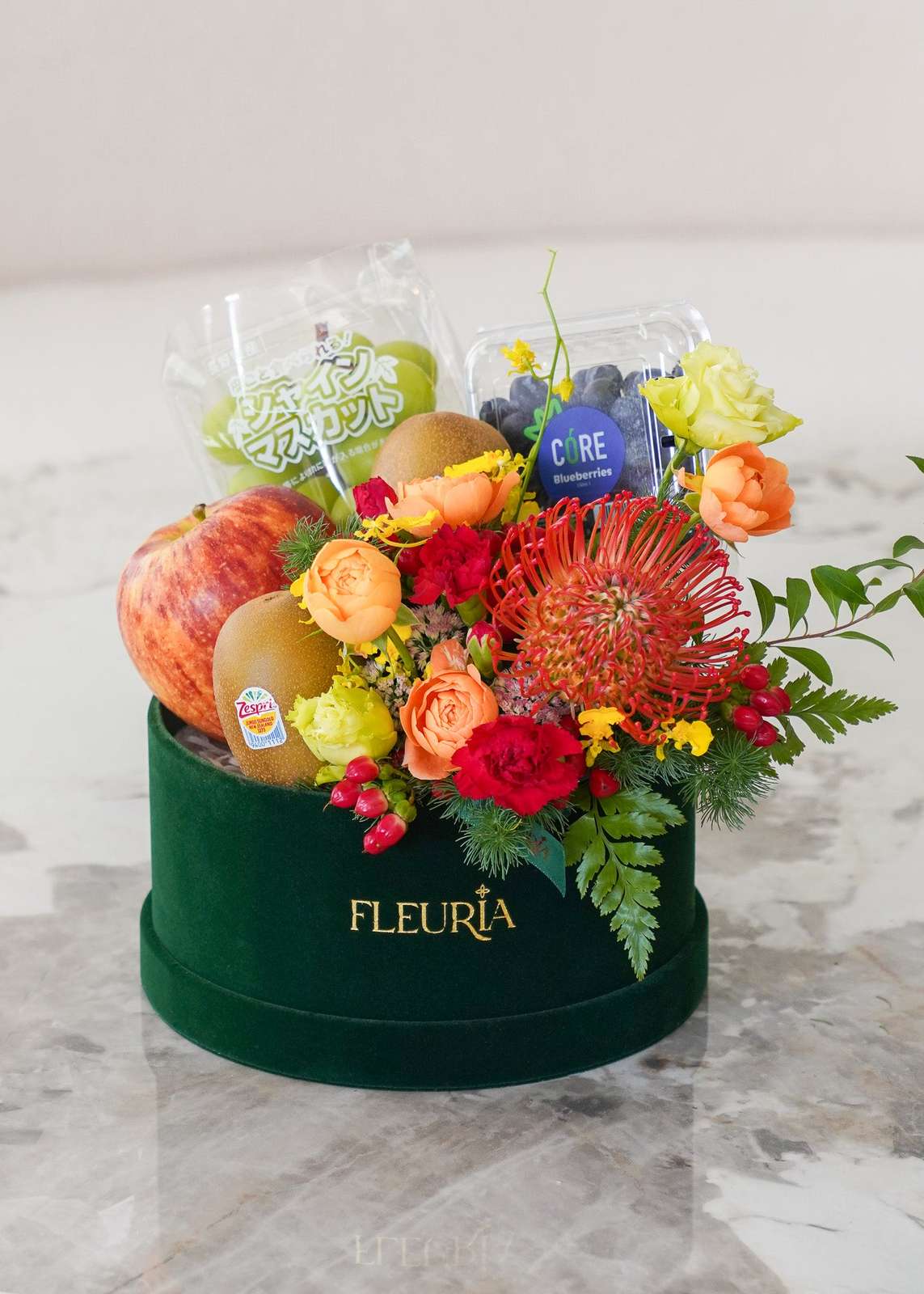 A round green velvet gift box with gold 'FLEURIA' branding, filled with a colorful fruit and flower arrangement. The bouquet