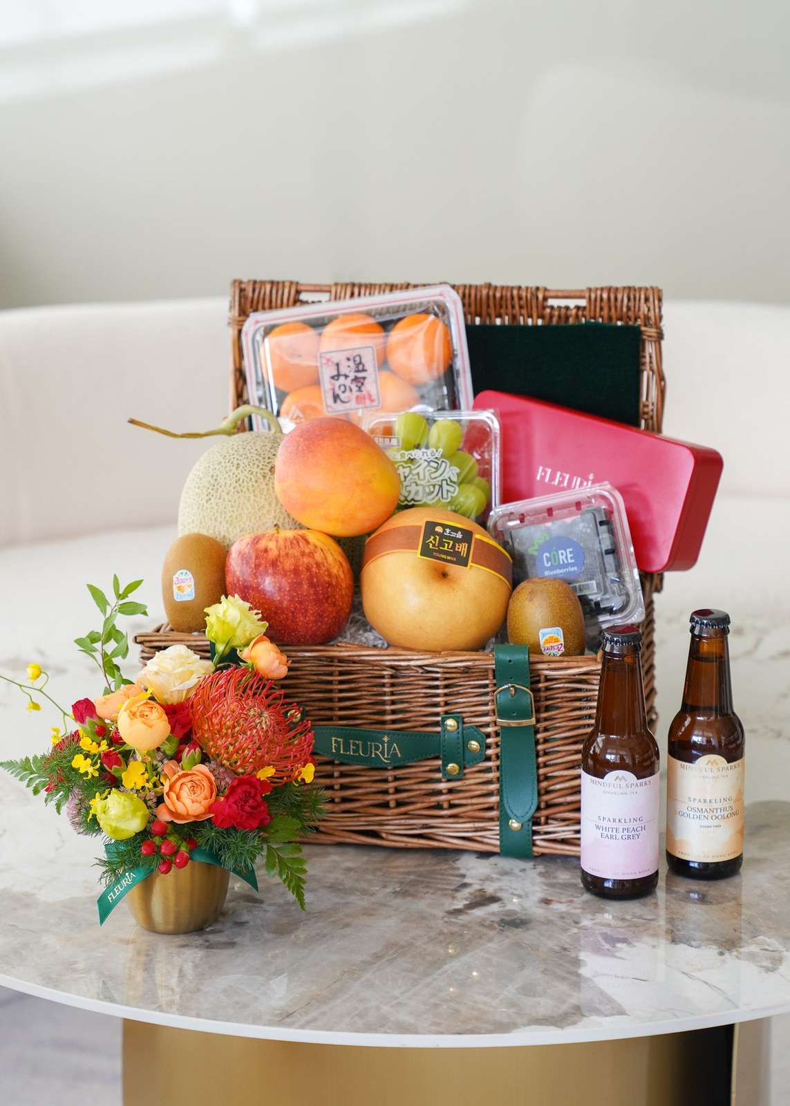 A wicker gift basket filled with assorted fruits, packaged vegetables, and a pink box, styled on a marble table with a small