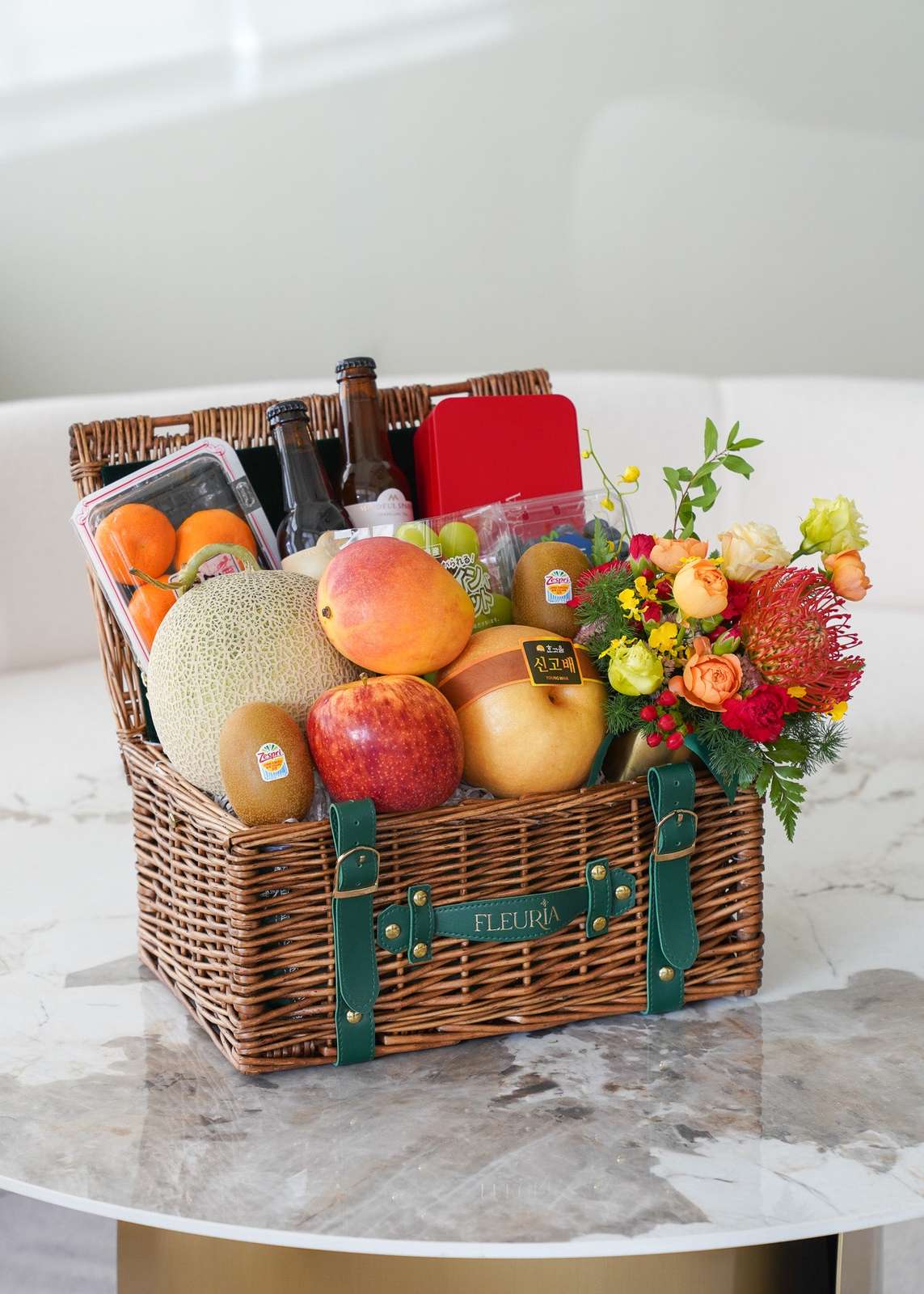 A wicker picnic basket filled with assorted fruits, vegetables, a bottle, and a colorful flower bouquet, displayed on a marbl