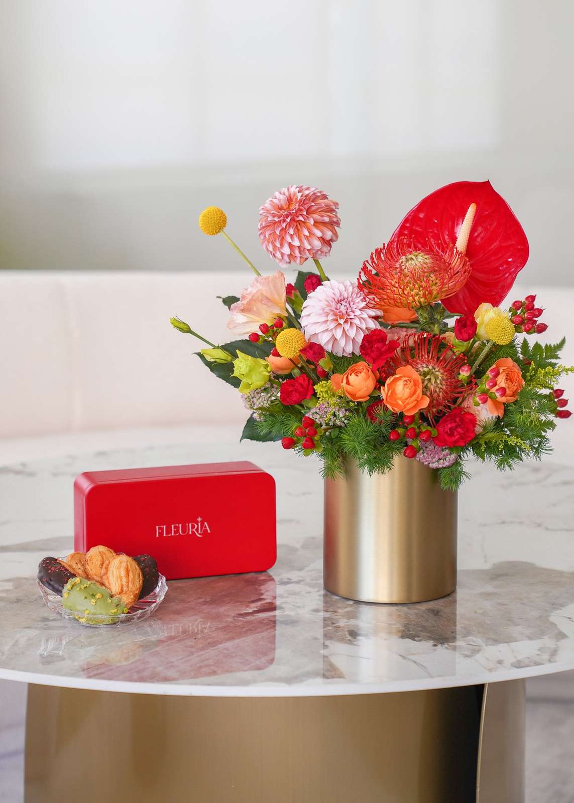 A vibrant floral arrangement in a metallic gold cylinder vase, displayed on a marble-topped table next to a red rectangular b