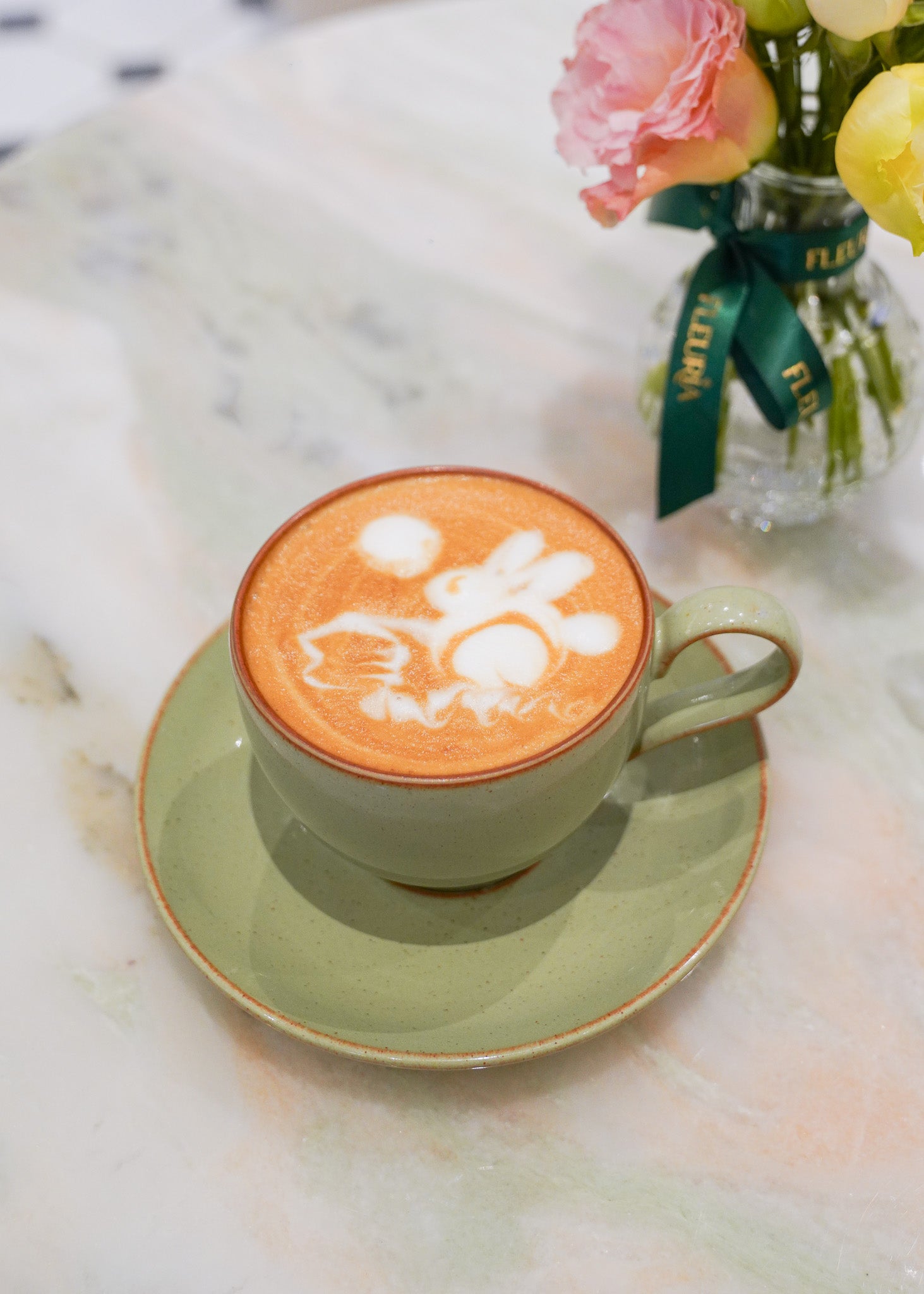 A light green ceramic cappuccino cup and saucer with a Jade Rabbit latte art design on top, set on a white marble table next to a small vase of pink and yellow flowers.