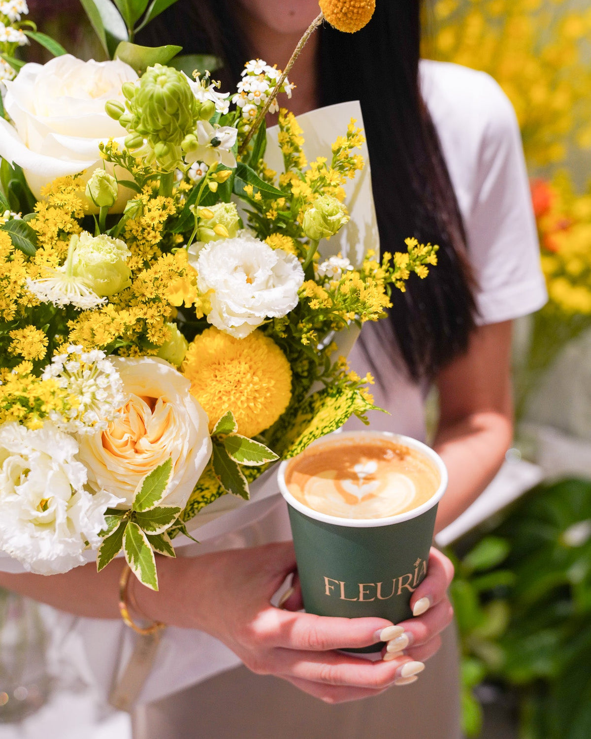A person holding a large bouquet of yellow and white flowers next to a green paper coffee cup printed with the brand name 'FLEURIL'.